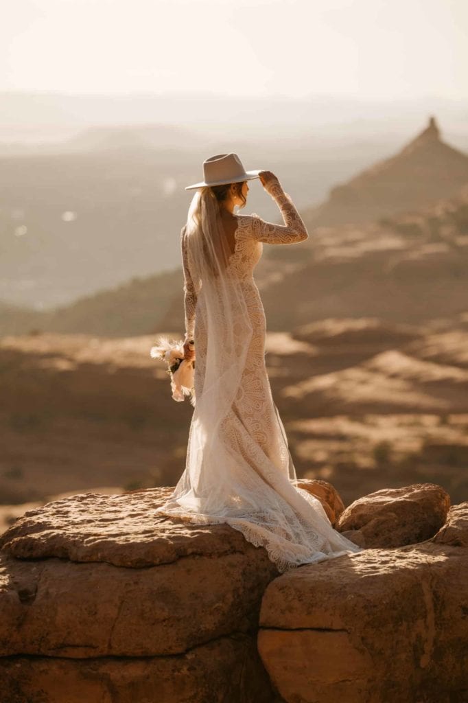 Bride in a wedding dress on an overlook, holding onto her bouquet and the tip of her hat.