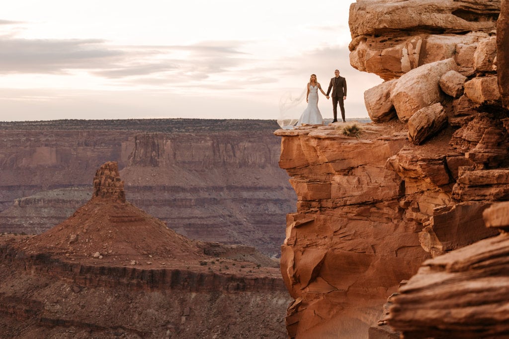 A couple stands holding hands at sunset on a red rock cliff overlooking the canyons. 