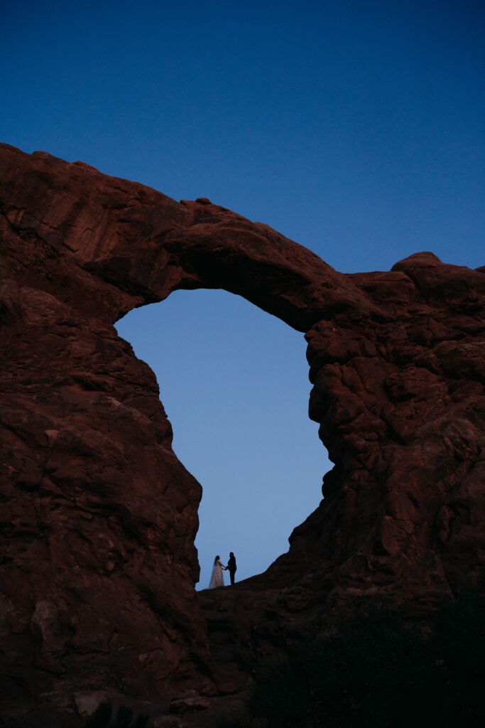 A couple stands in the center of an arch at dusk.
