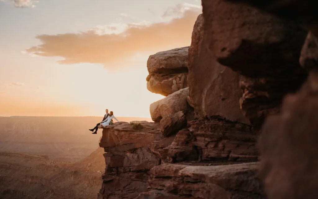 A couple sits on an edge of a cliff at sunset in Moab.