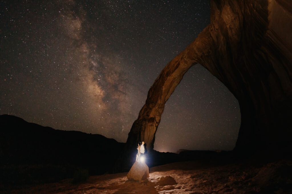 A couple stands on a rock in Moab under the Milky Way.