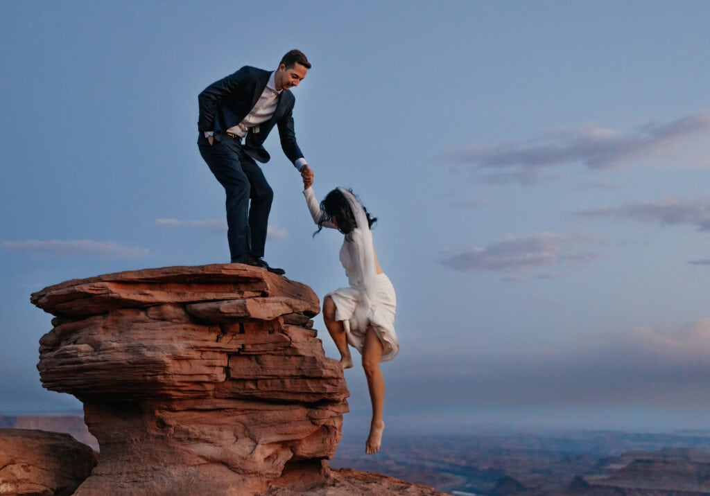 A groom helps a bride up a rock at dusk.