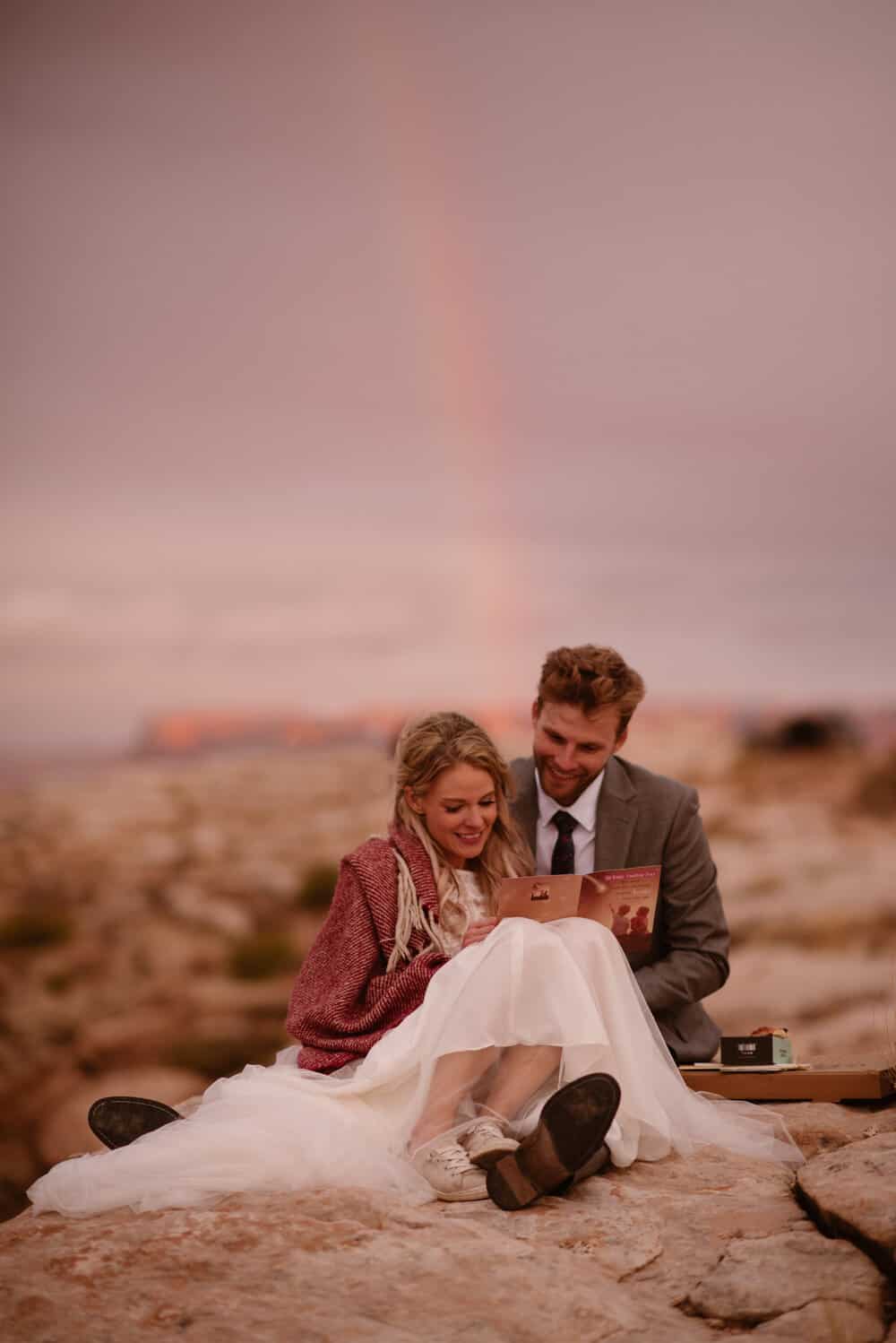 A bride and groom read letters together while a rainbow shines behind them.