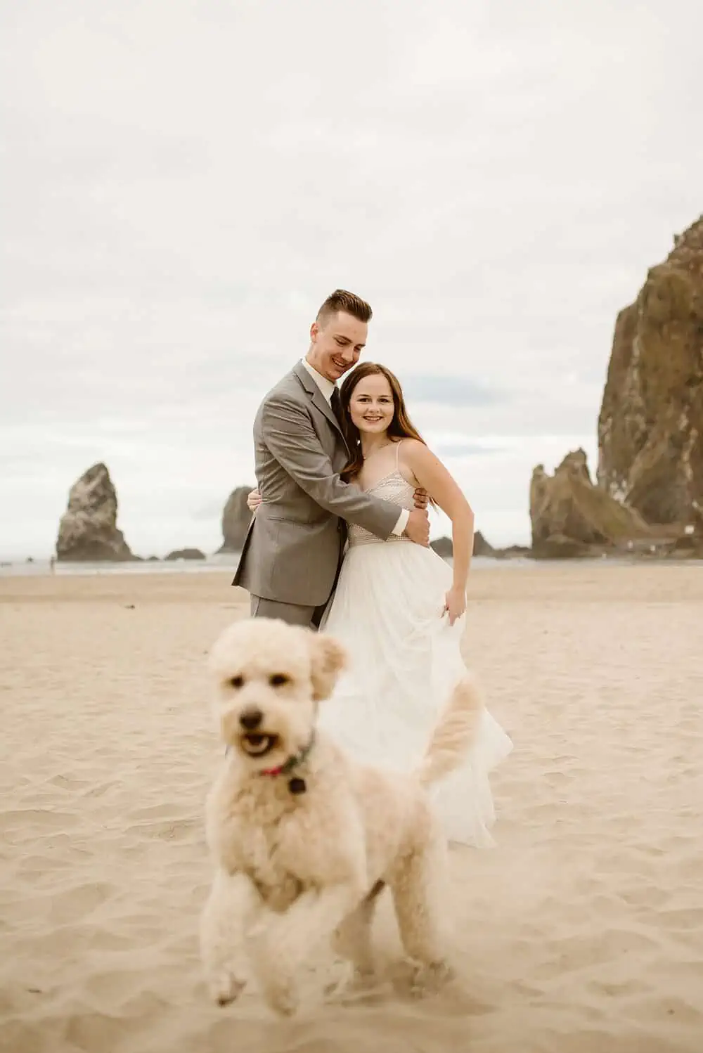 A couple smiles for a portraits as their dog runs toward the camera on the beach.