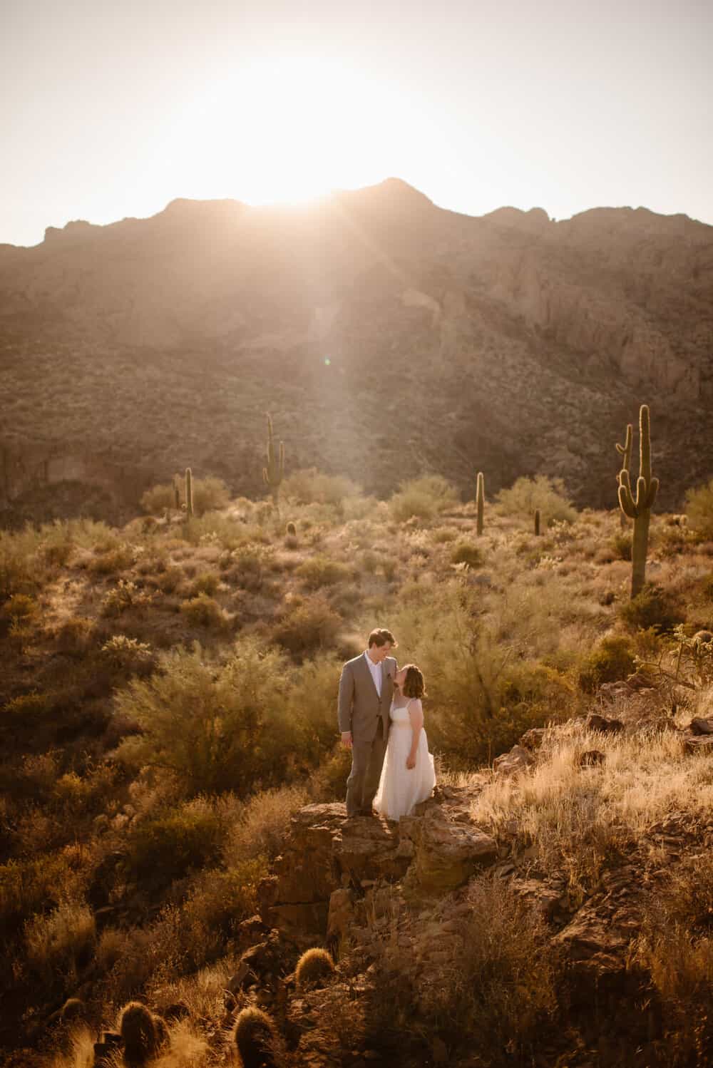 A couple stands in the desert together as the sun sets behind them.