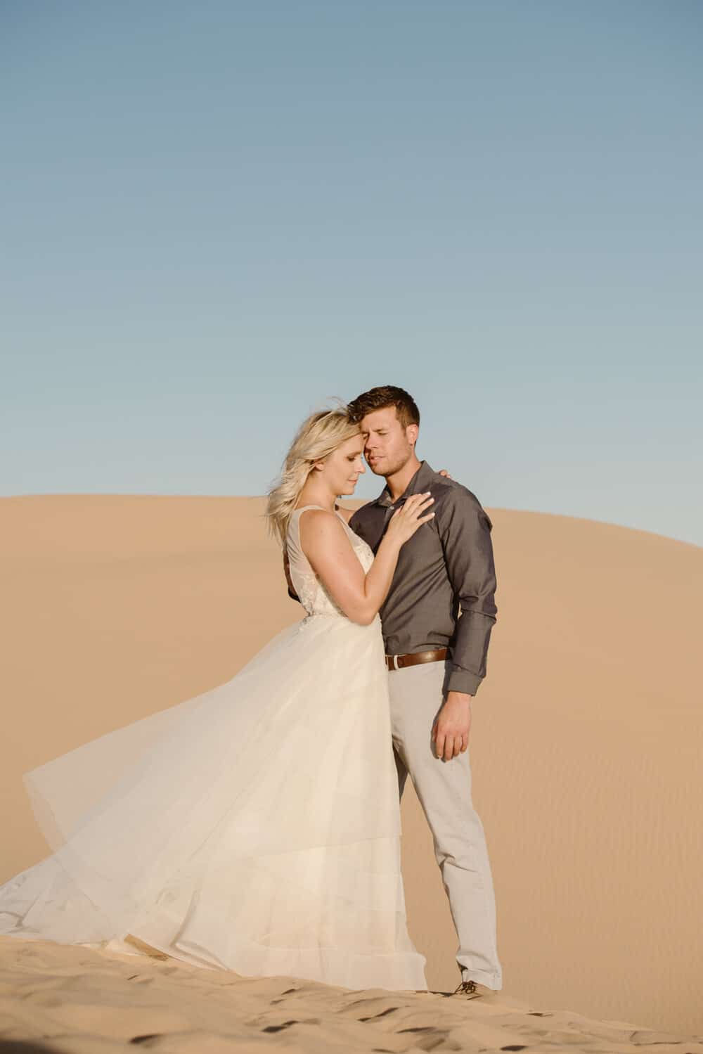A bride and groom stand together on a sand dune in the middle of the day.