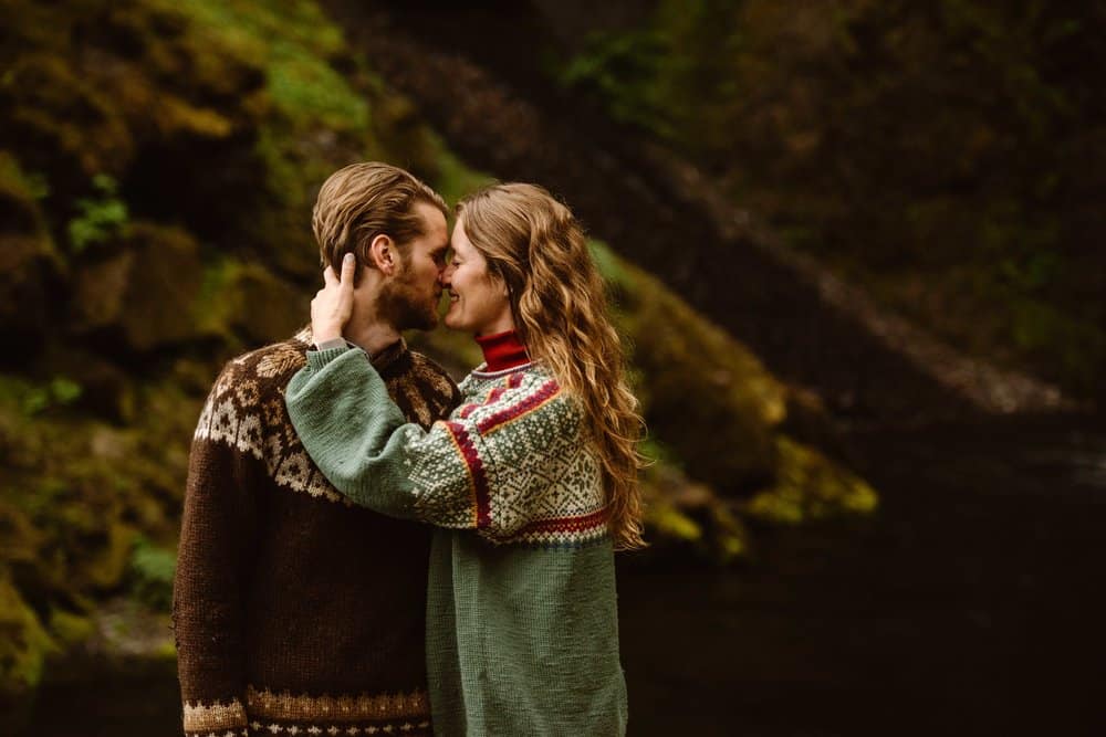 A couple in Nordic sweaters standing in front of a mossy wall together. 
