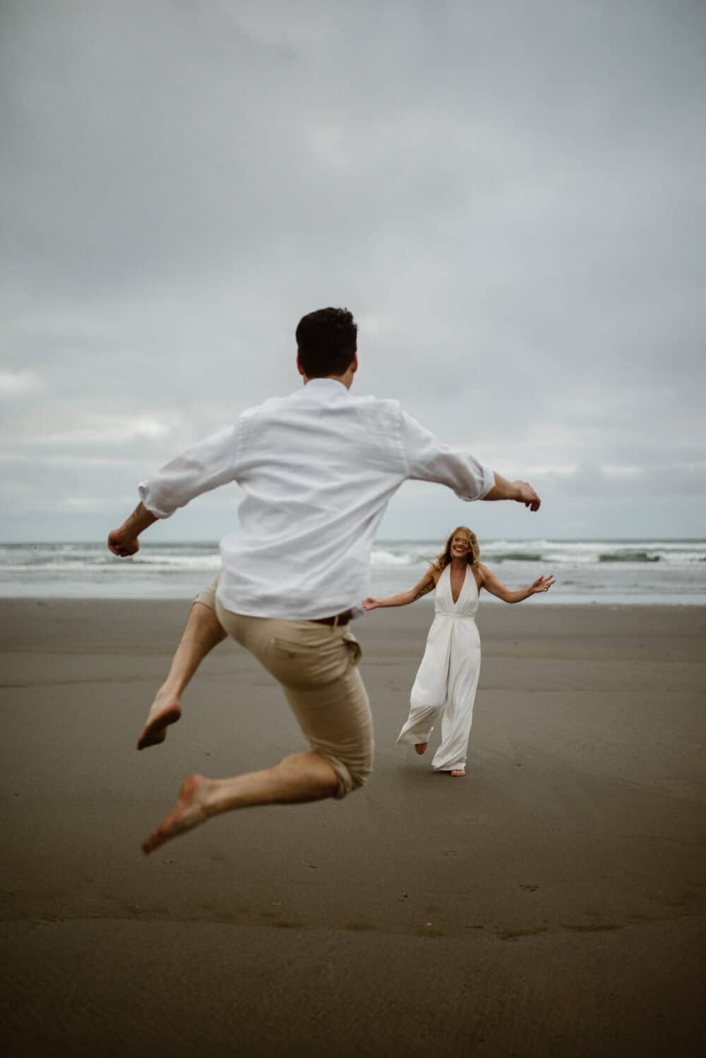 A groom jumps for joy on the beach running towards his bride. 