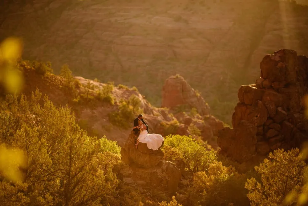 A man and woman sit on a rock together looking at the Rock they just climbed as the sun sets behind them.