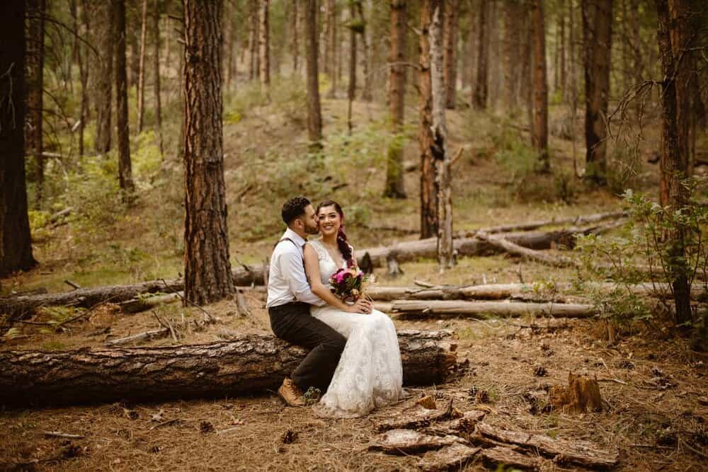 A groom kisses his bride on the cheek as they sit on a log.