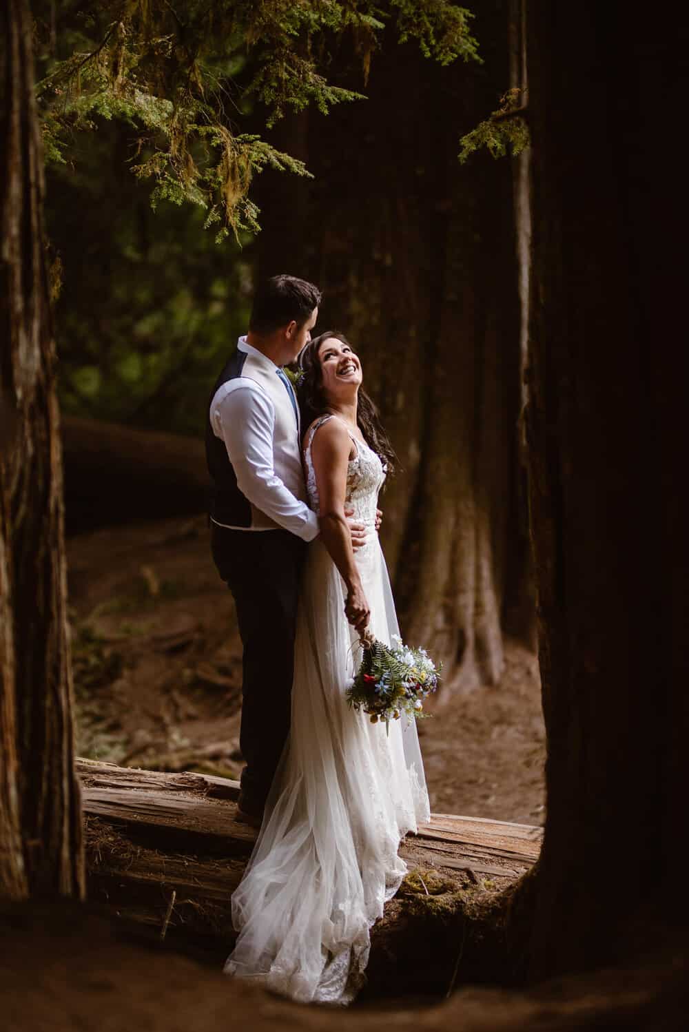 A bride smiles at her groom as they stand together in a forest. 