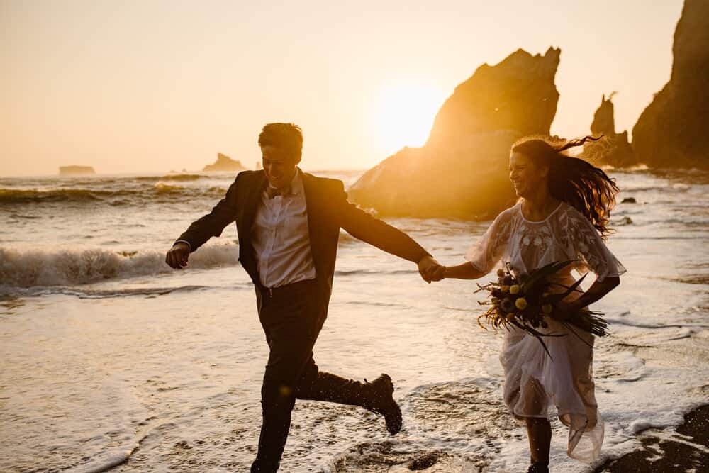 A bride and groom run through the water as the sun sets behind a rock behind them and the waves crash in toward them.