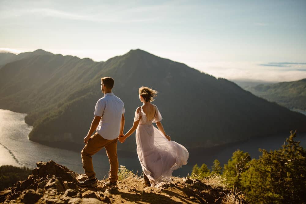 A man and woman stand facing the sun together overlooking a large body of water and mountain peak. 