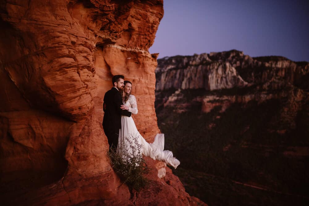 A couple stands on a red rock ledge after sunset.