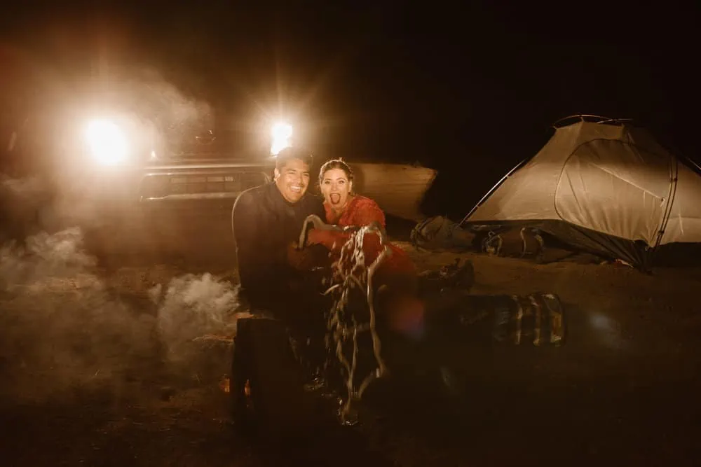 A couple smiles as they pop a bottle of champagne at their camp site lit up by headlights of their truck. 