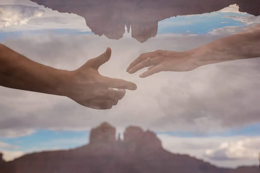 A couple reaching their hands towards each other with cathedral rock in the background.