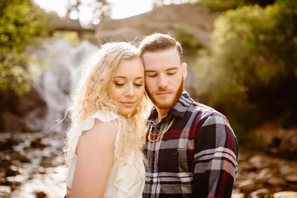 A couple hold their faces together while looking down towards something out of the frame while standing in an out of focus creek bed.