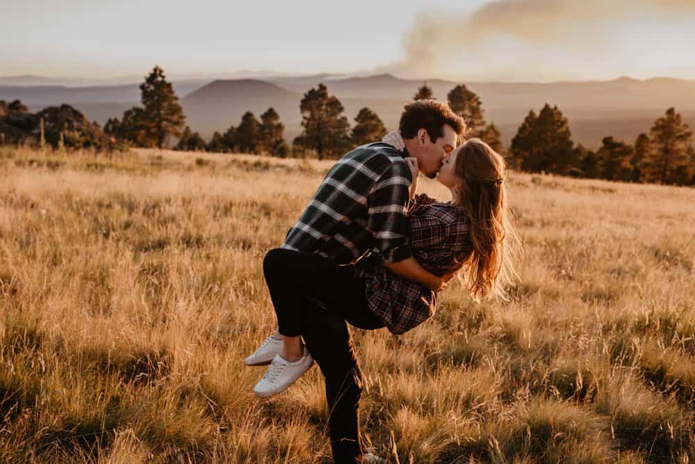 A man holds his woman in a field with smoke from a fire in the distant background and they share a kiss while he dips her back.