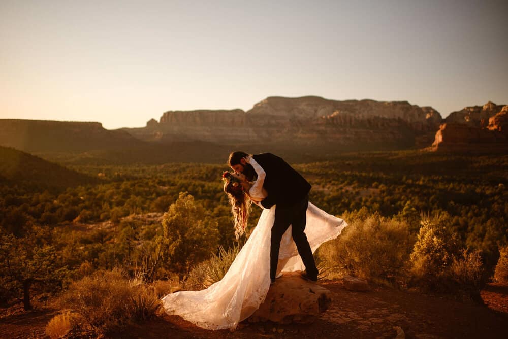 A groom dip kisses his bride surrounded by desert views at sunset.