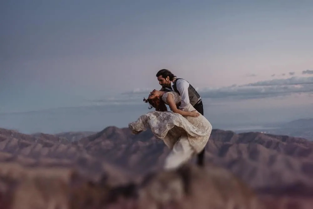 A groom dips his bride as her dress catches the wind as the last bit of light is left at the top of a mountain.