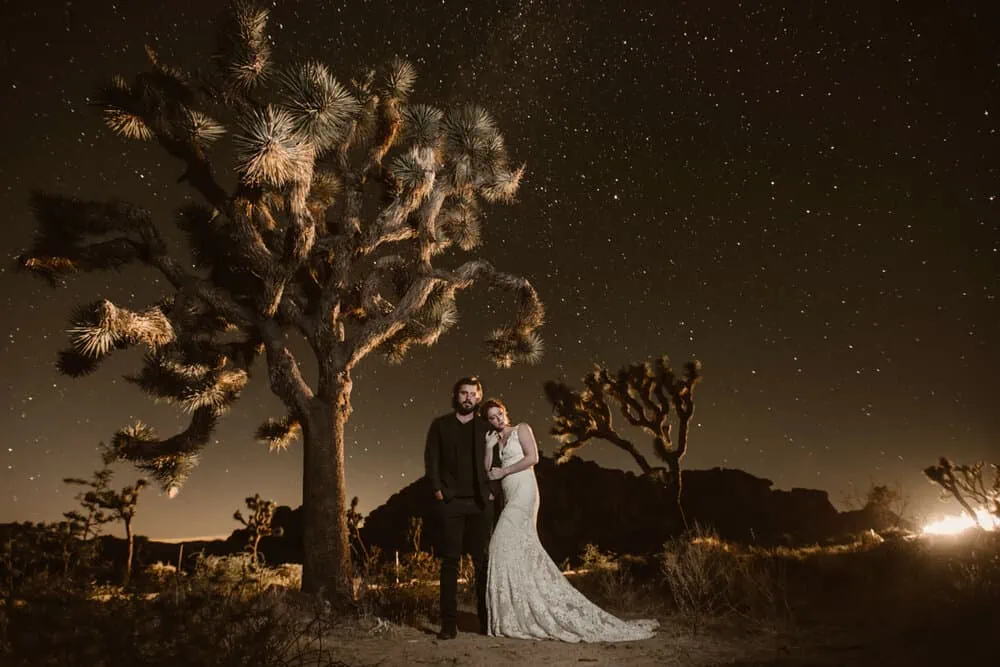 A couple holds each other close under a joshua tree and the stars as a car drives by.