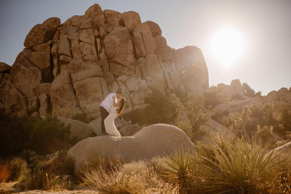 A couple share a kiss in the sun while standing on a boulder in front of a pile of rocks in Joshua Tree.
