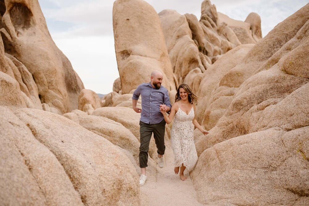 A couple runs through the rocks of Joshua Tree together smiling ear to ear. 