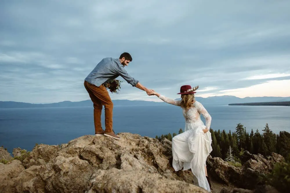 A groom helps a bride up a rocky cliff.