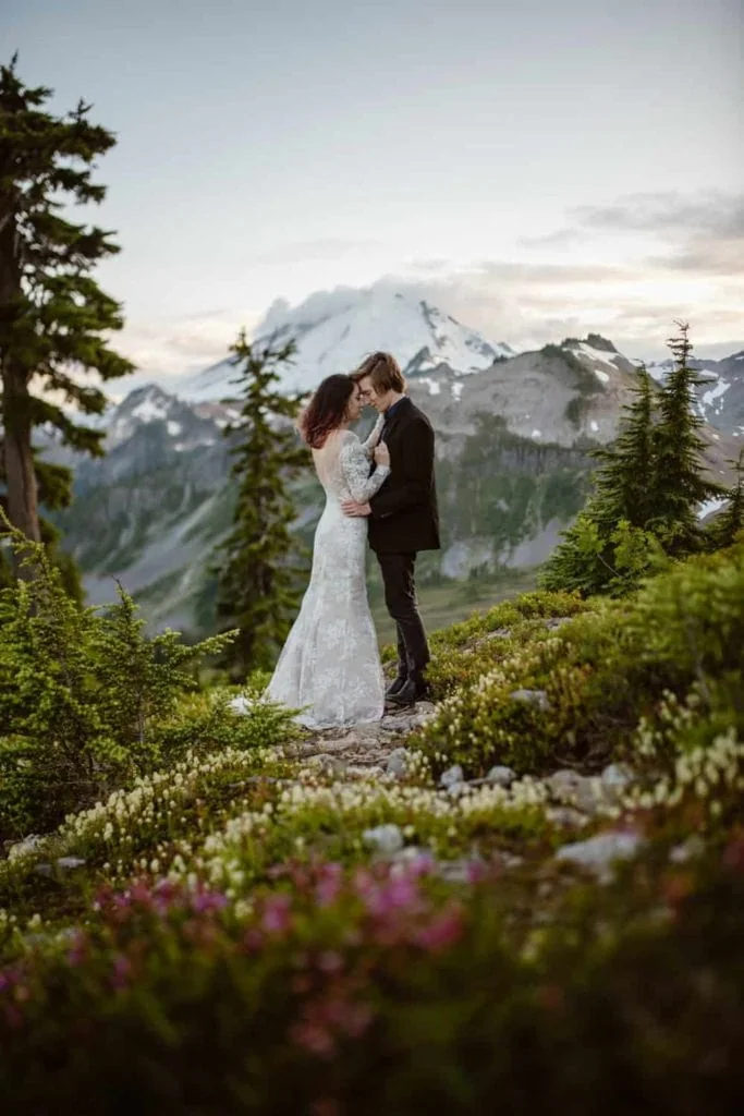 A couple holds each other close at dusk as wildflowers and mountains surround them.