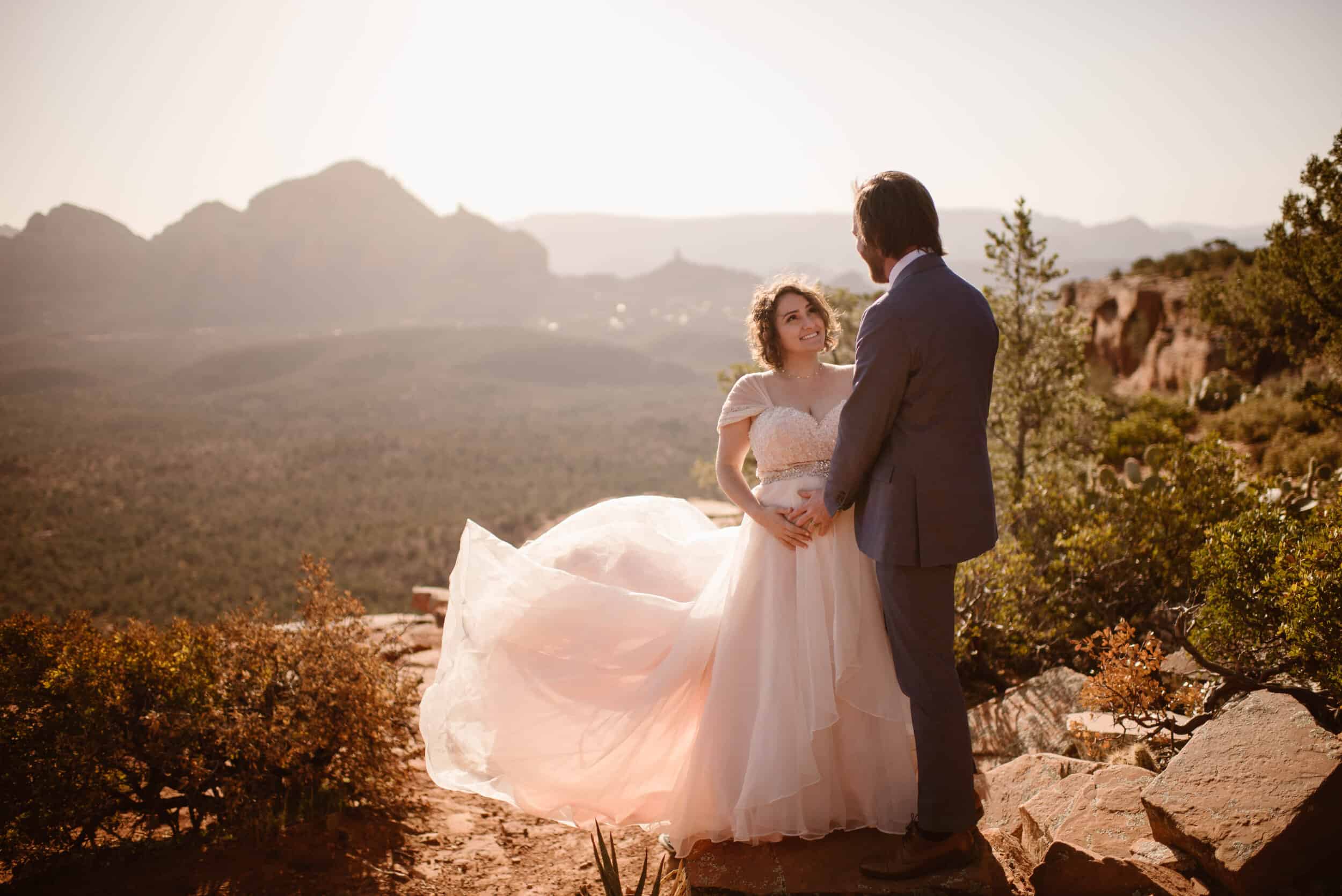A pregnant bride and groom stand together as the sun rises over the red rocks of Sedona Arizona.