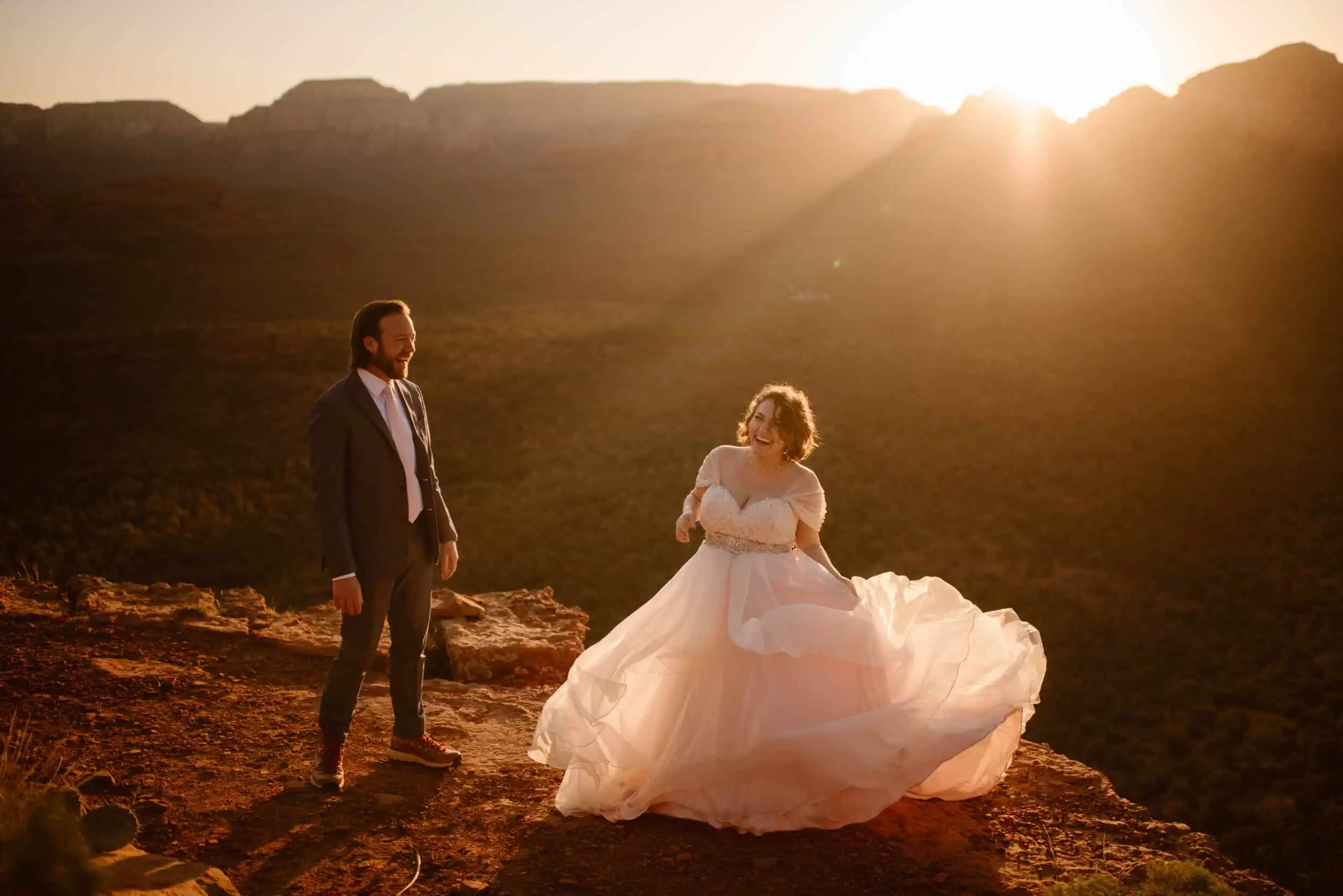 A groom and his pregnant bride stand together on the edge of the Sedona Red Rocks while the sun rises over the mountains.