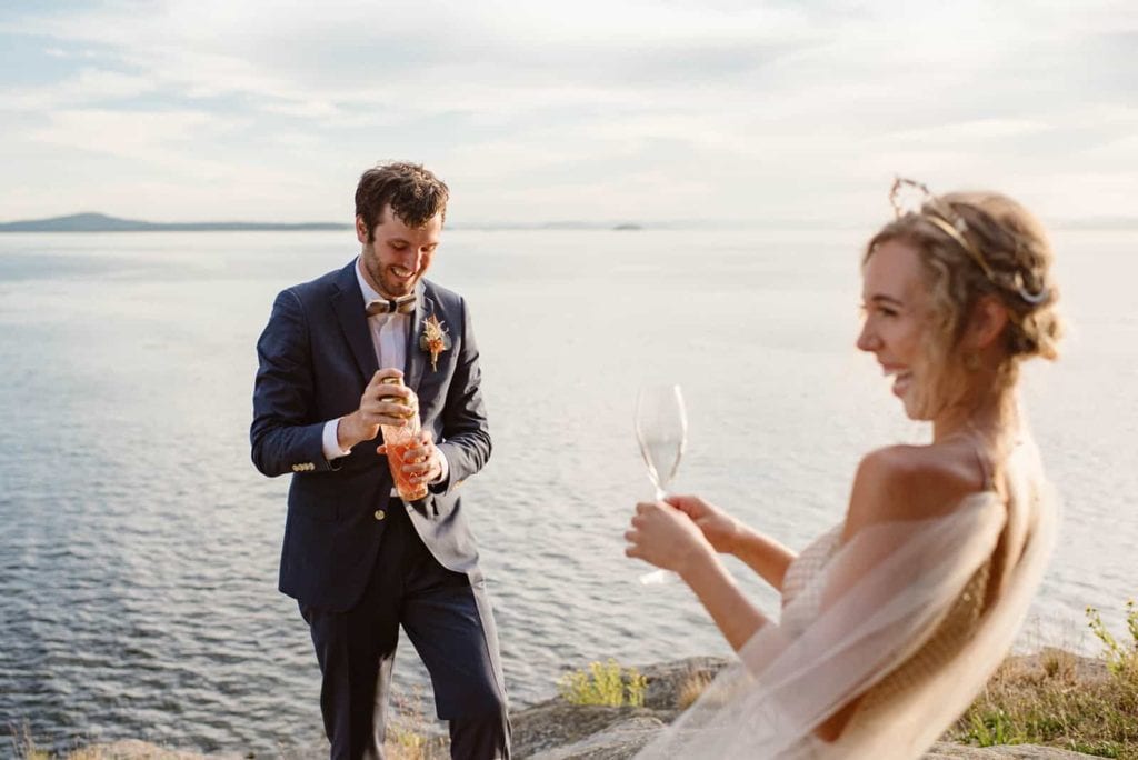 A bride and groom laughing as the groom mixes a cocktail while she holds a glass right on the coast.