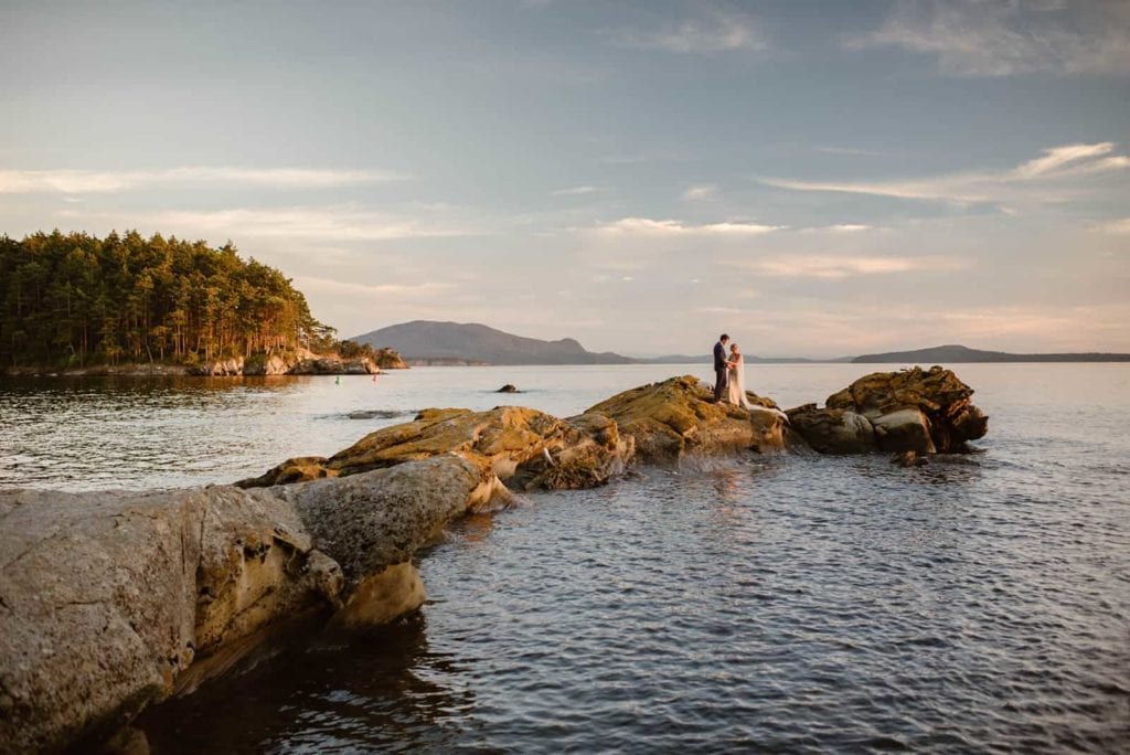 A bride and groom stand together on a distant point at the edge of Sucia island.