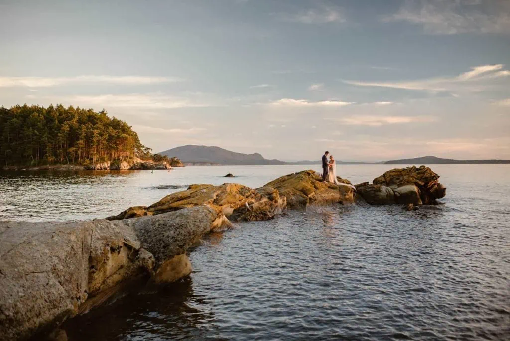 A bride and groom stand together on a distant point at the edge of Sucia island.