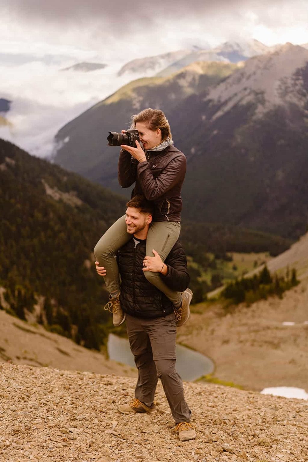 A man holds a woman on his shoulders as she photographs something while mountain views are visible behind them.