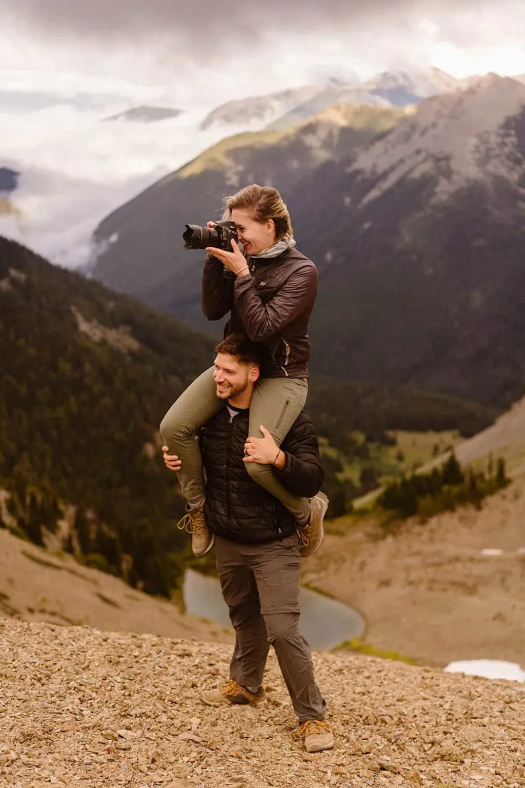 A photographer sits on a mans shoulders to take a photo in the mountains. 
