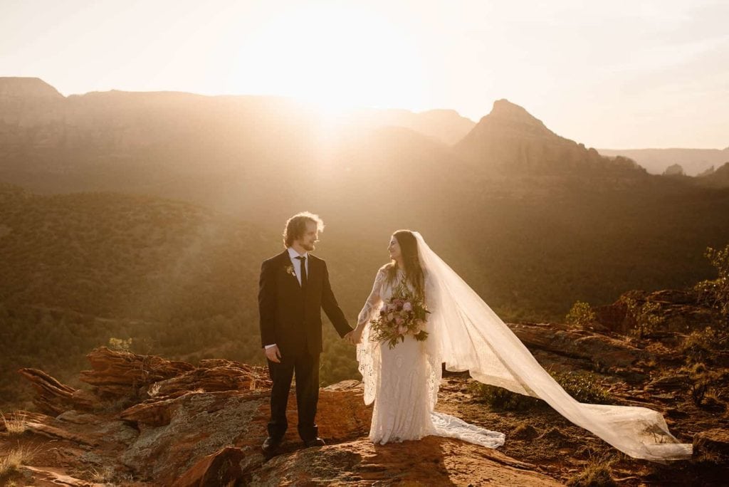 The bride and groom stand together holding hands as the sun rises over Sedona.