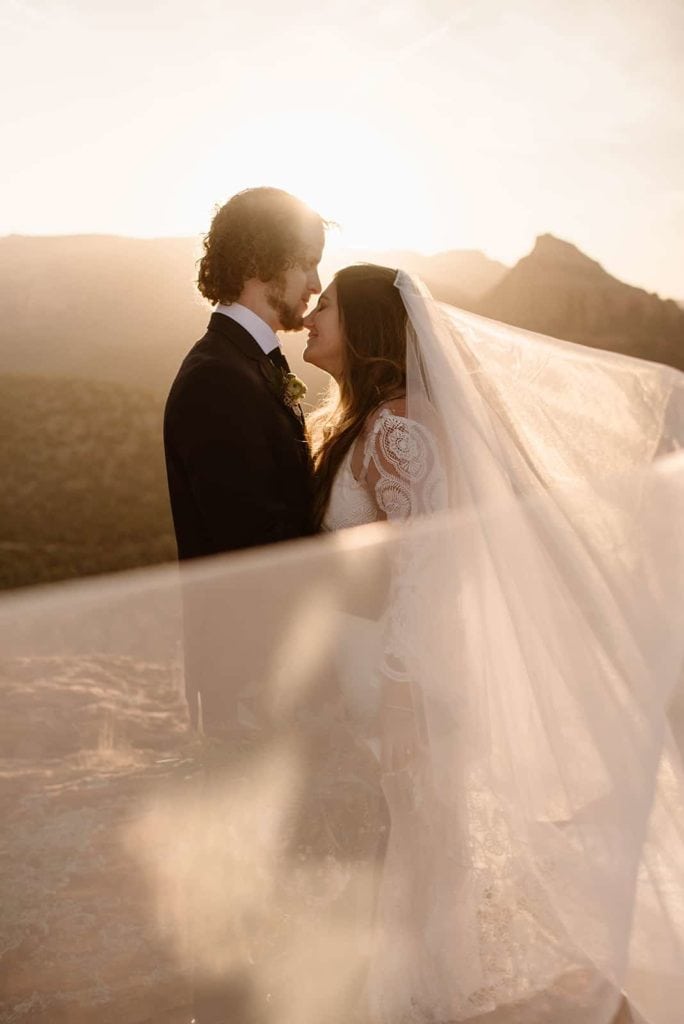 The brides veil blows in the wind while she stands with her groom at sunrise in Sedona