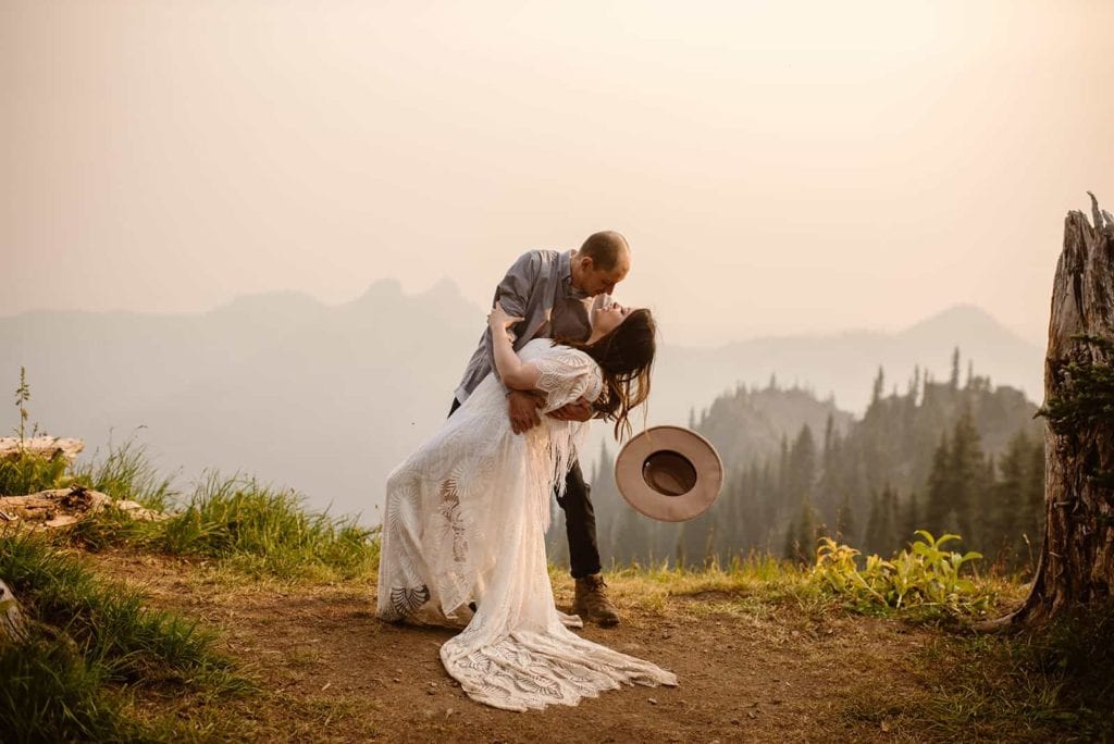 A man dips his bride to give her a kiss while her hat falls off and smoke fills the morning air at sunrise.