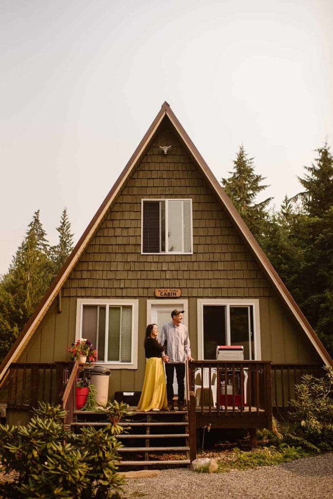 A man and his wife stand together on the front porch of an a frame cabin surrounded by trees.