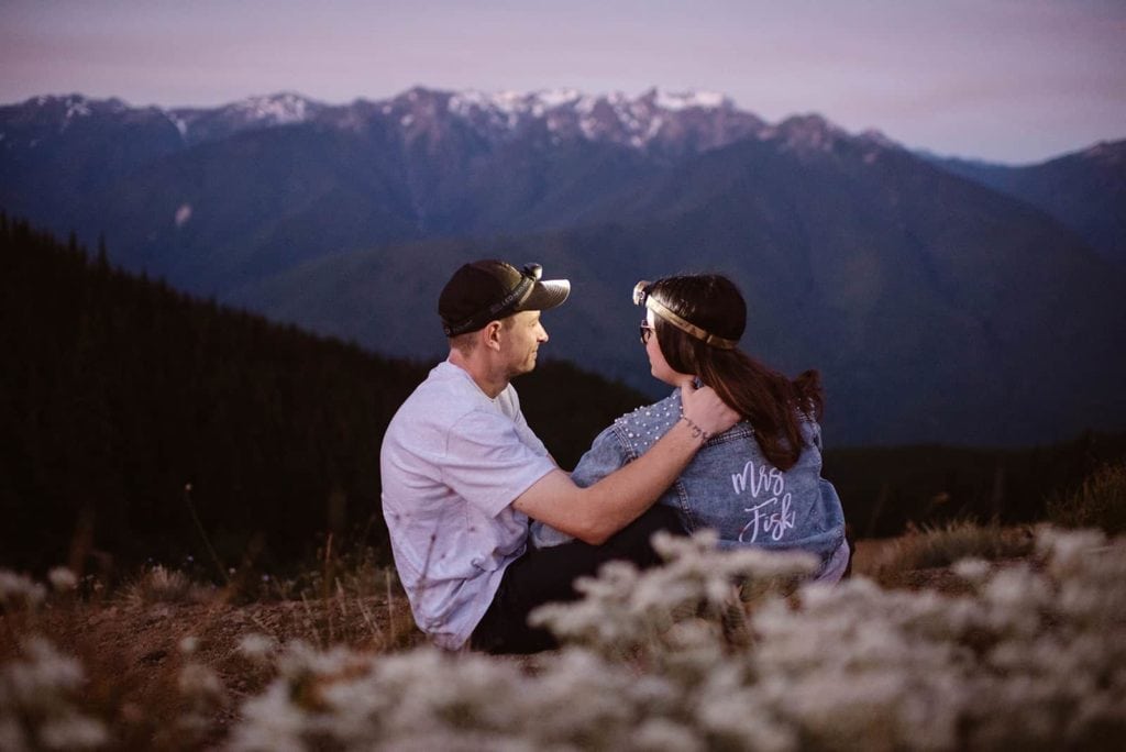 A man and his fiancee sitting together on the edge of a cliff with mountain views in the distance.
