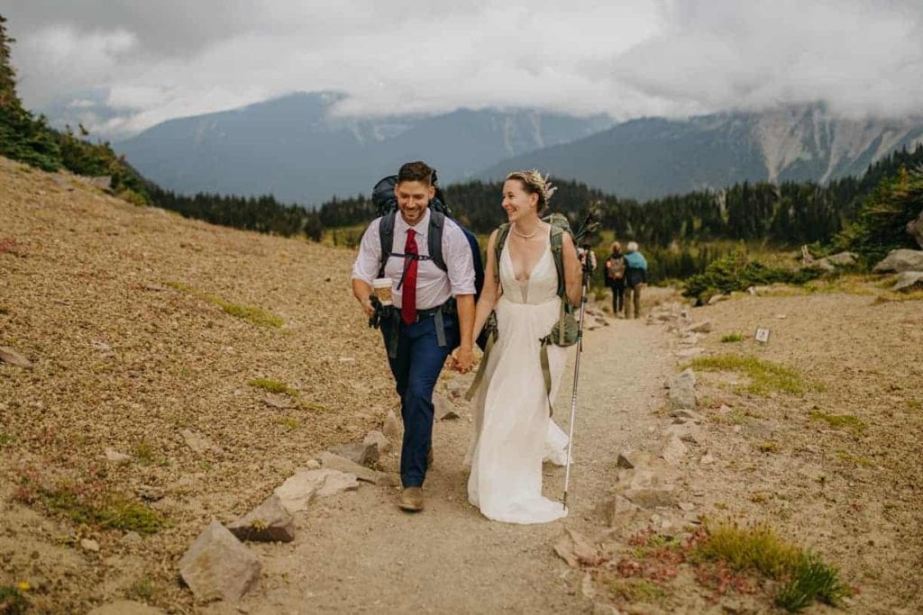 A Bride and groom walk together in wedding clothes in Mount Rainier on a trail.