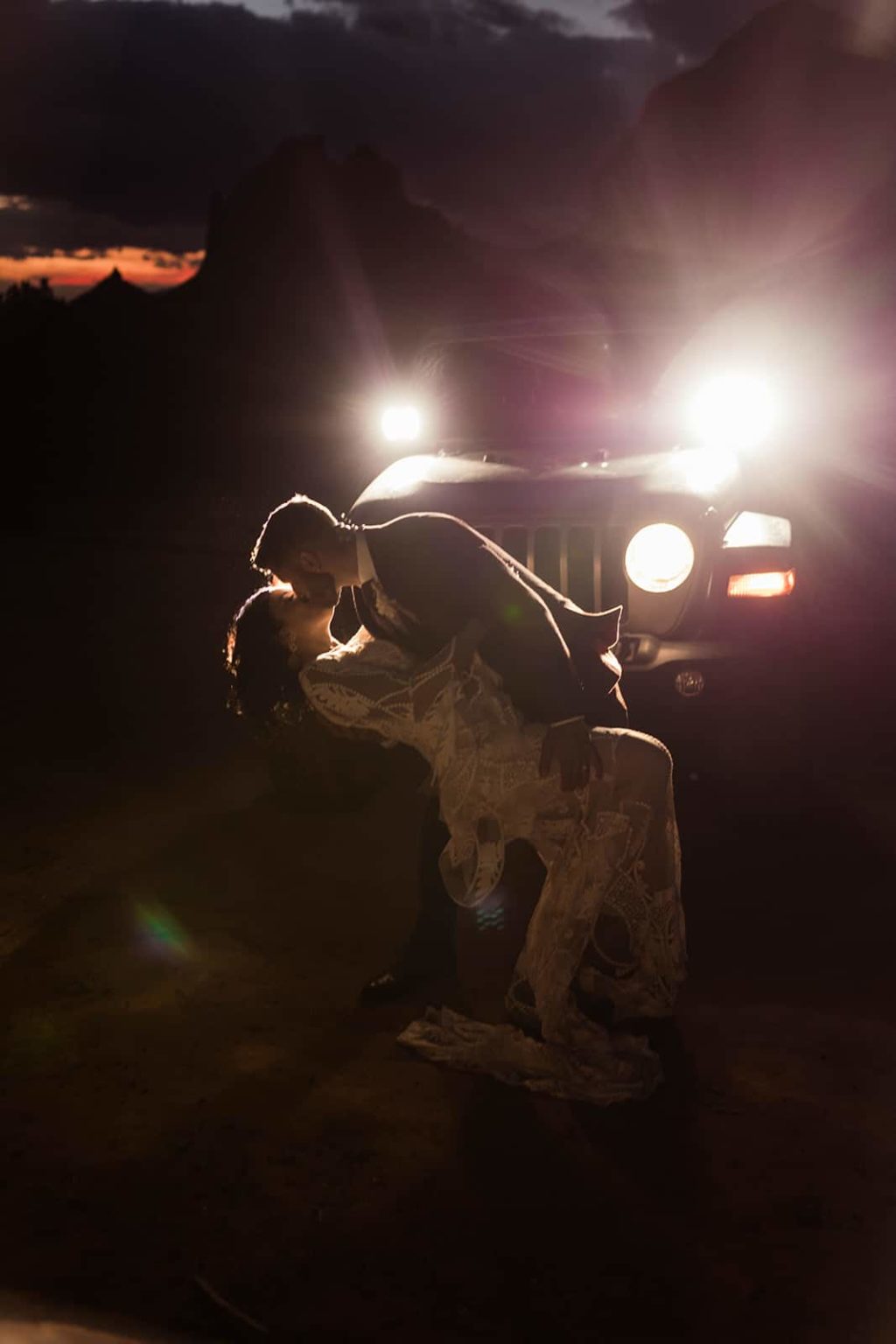 The groom dip kisses his bride in front of the lights of their lit up jeep with the last bit of light in the Sedona skies.