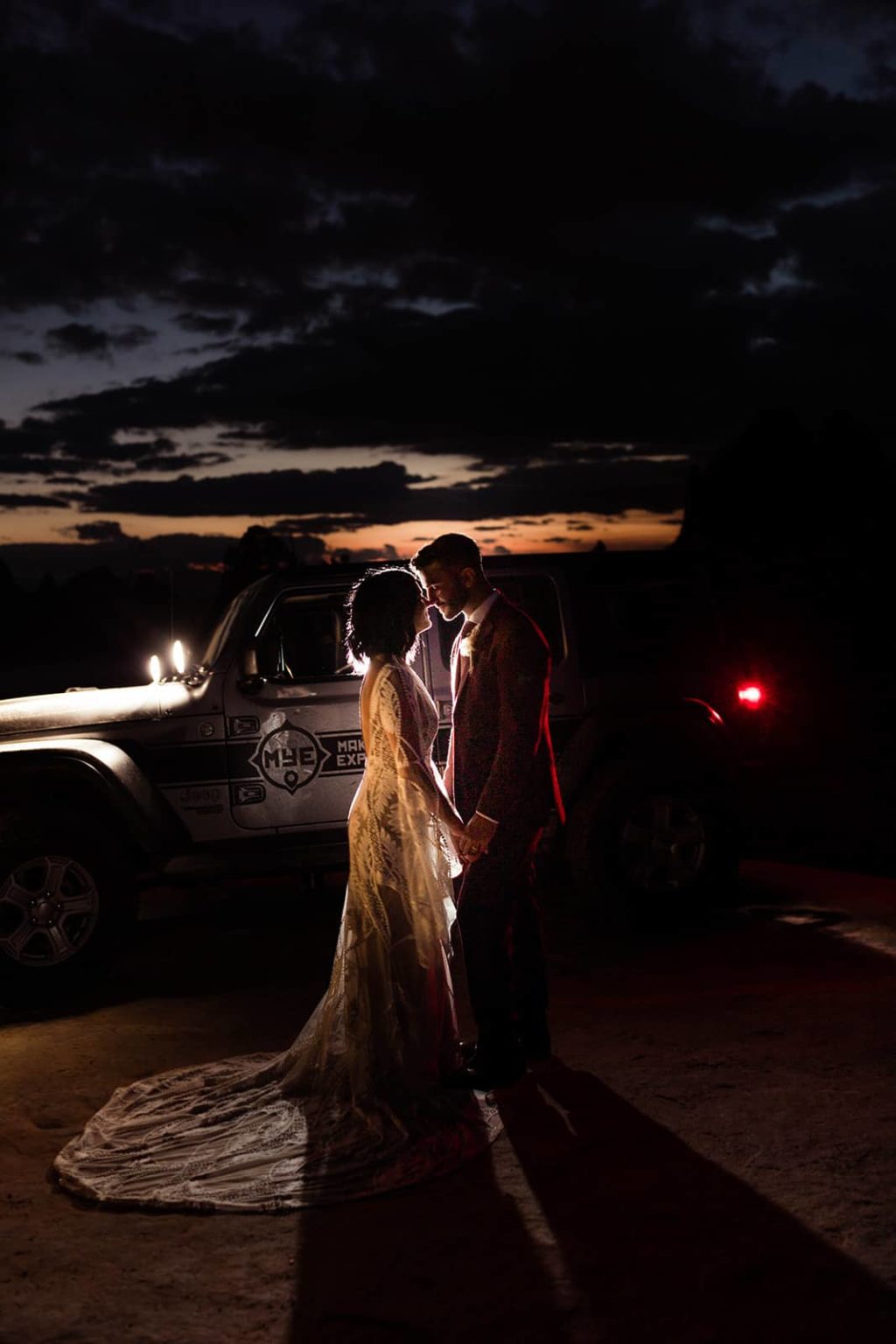 The bride and groom stand close together with the little light left in the sky and their jeep in the background