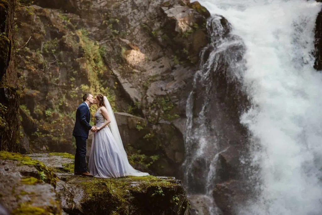 A bride and groom share a kiss while standing near a waterfall.