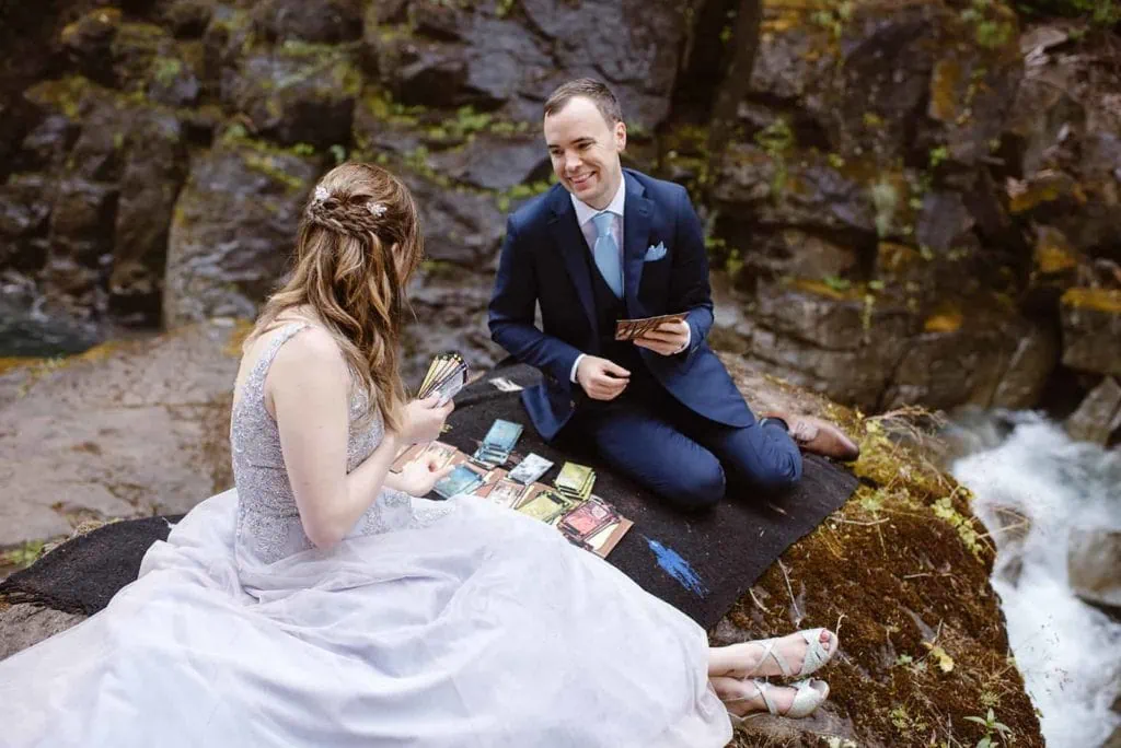 A bride and groom play a card game together near a waterfall.