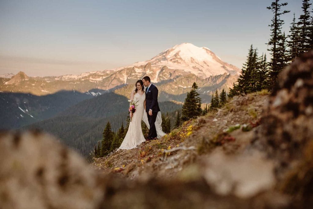 A couple shares a private moment together standing on a mountains side in the. morning light with mount rainier behind them.