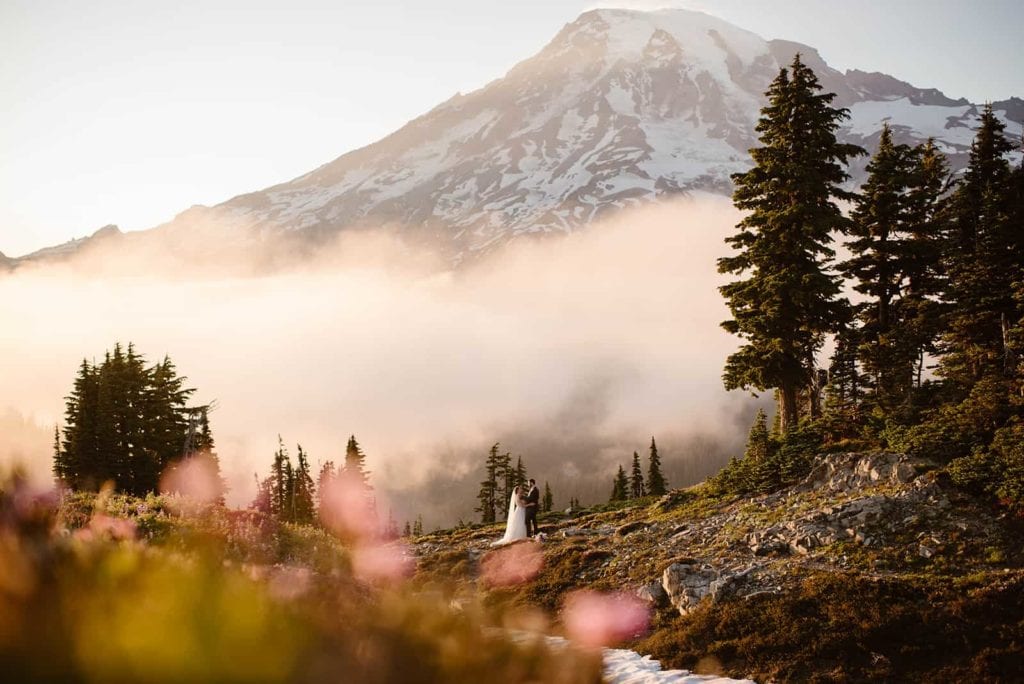 A bride and Groom hold each other close on a trail overlooking Mount Rainier with wildflowers in the foreground.