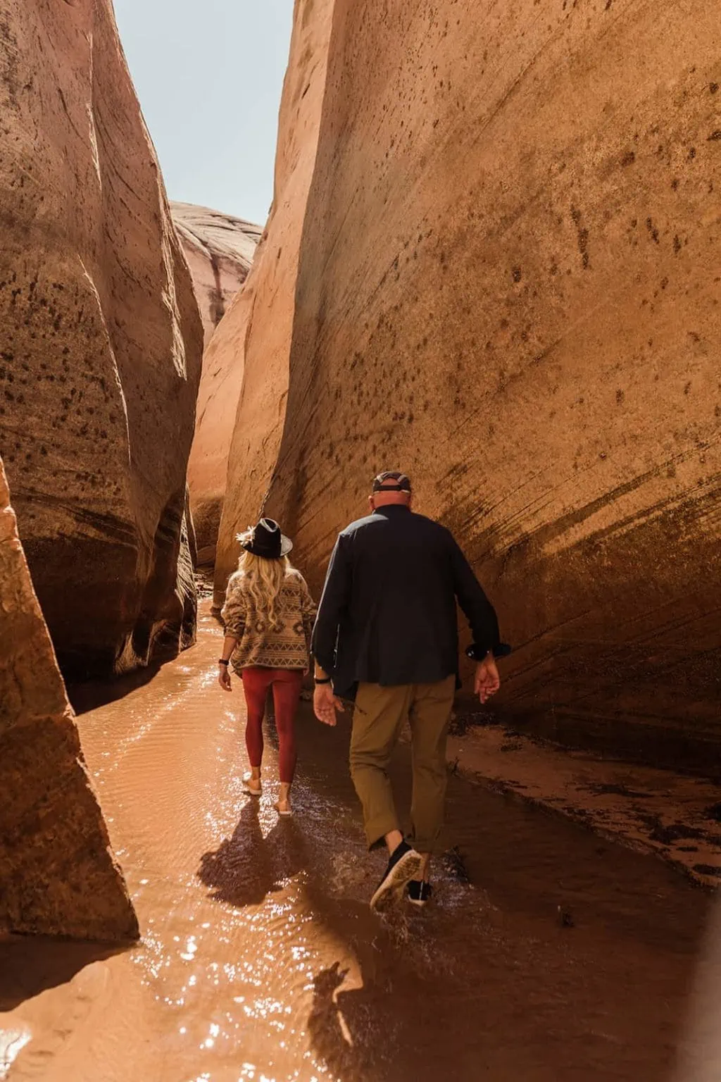 A couple walks together through water in a red rock slot canyon.