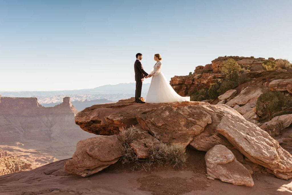 A couple holds hands and looks into each others eyes as they stand on a sandstone boulder overlooking Moab. 
